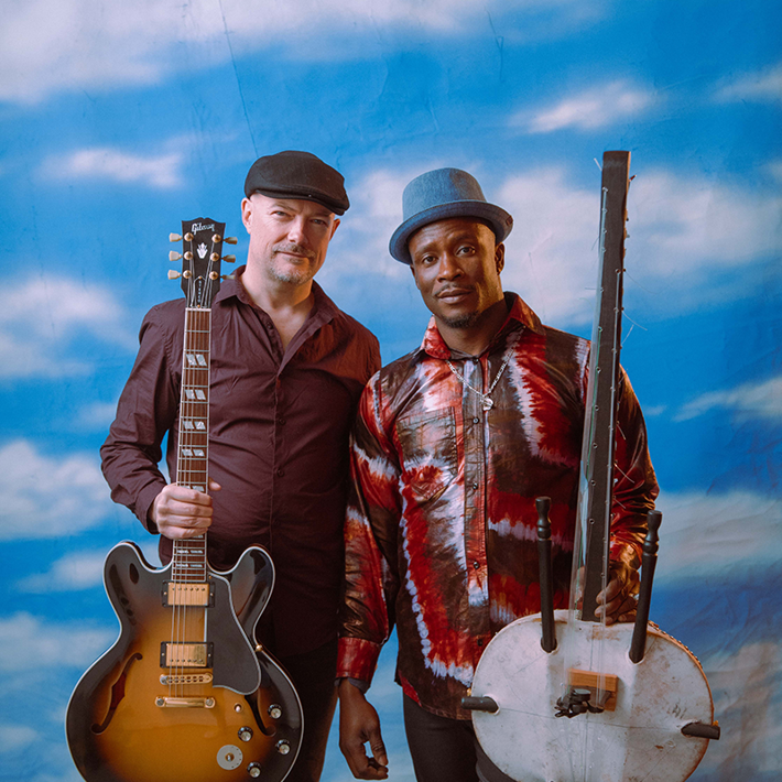 Two men wearing hats stand facing the camera, in front of a backdrop with blue skies and clouds. The men are holding a guitar and a kora (African harp).