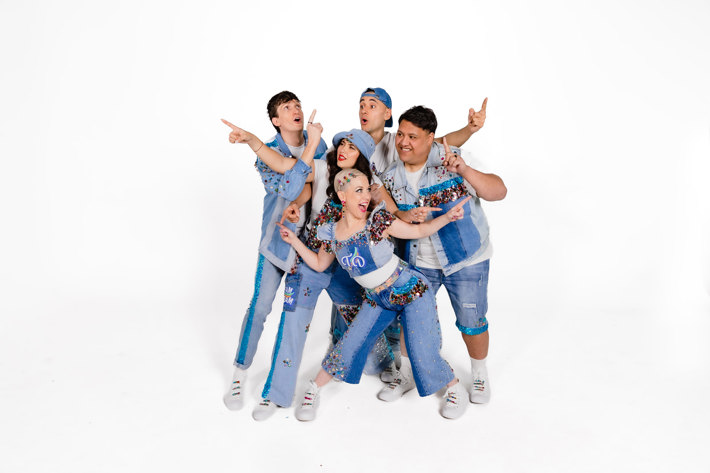 Group of five band members in bejewelled denim outfits posing energetically against a white background.
