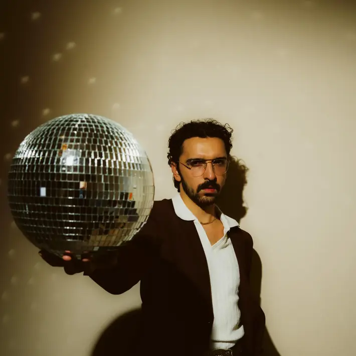 Ricky Neil Jr, a man with black hair and dark moustache, wearing a dark suit with white shirt and large collar. His right arm outstretch in front of his body, holding a disco ball, his left hand in his pocket