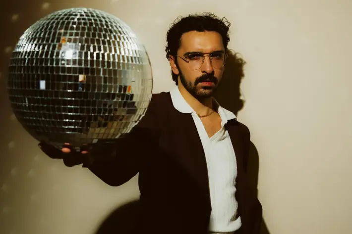Ricky Neil Jr, a man with black hair and dark moustache, wearing a dark suit with white shirt and large collar. His right arm outstretch in front of his body, holding a disco ball, his left hand in his pocket