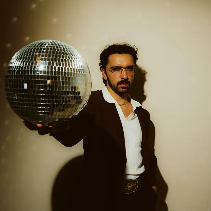 Ricky Neil Jr, a man with black hair and dark moustache, wearing a dark suit with white shirt and large collar. His right arm outstretch in front of his body, holding a disco ball, his left hand in his pocket