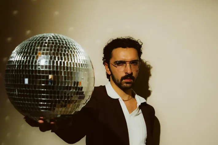 Ricky Neil Jr, a man with black hair and dark moustache, wearing a dark suit with white shirt and large collar. His right arm outstretch in front of his body, holding a disco ball, his left hand in his pocket