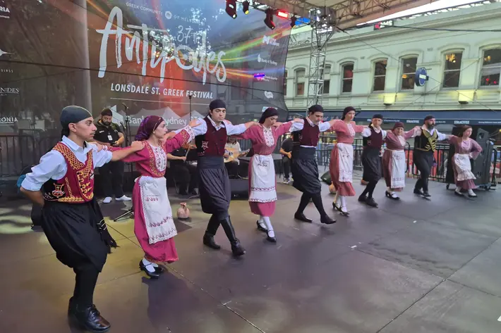 A group of performers in traditional Greek attire dance on an outdoor stage at the Lonsdale Street Greek Festival. They are dressed in coordinated costumes featuring embroidered vests, aprons, and skirts, with black shoes and headpieces. The dancers are linked arm-in-arm in a line formation, while musicians play instruments in the background. A large banner reading “Antipodes” is displayed behind them, with colourful stage lighting and historic buildings visible beyond the stage.