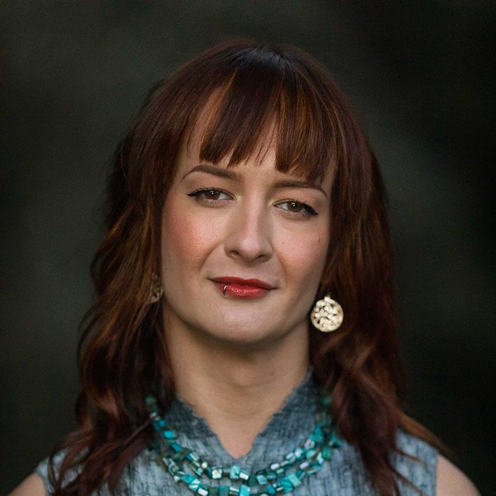 Celeste, a person with long brown hair wearing a gray textured top with a necklace of turquoise stones. They have a serious, thoughtful expression on their face and are looking directly at the camera.