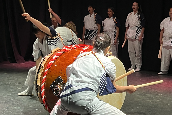 A group of performers is engaged in a traditional Japanese Taiko drumming performance on stage. Two large drums with decorative ropes and detailed designs are positioned in the foreground, with performers striking them using wooden drumsticks in dynamic stances. Other participants stand in the background holding drumsticks, dressed in coordinated white and blue attire. The setting is indoors with a dark backdrop, highlighting the vibrant energy and cultural significance of the performance.
