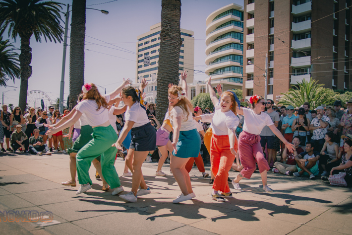 A group of people dancing outdoors in colorful 80s style clothing with an audience watching, set against a backdrop of palm trees and tall buildings.