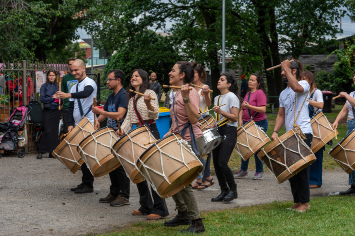 A group of people playing large drums outdoors, surrounded by trees and a small crowd.