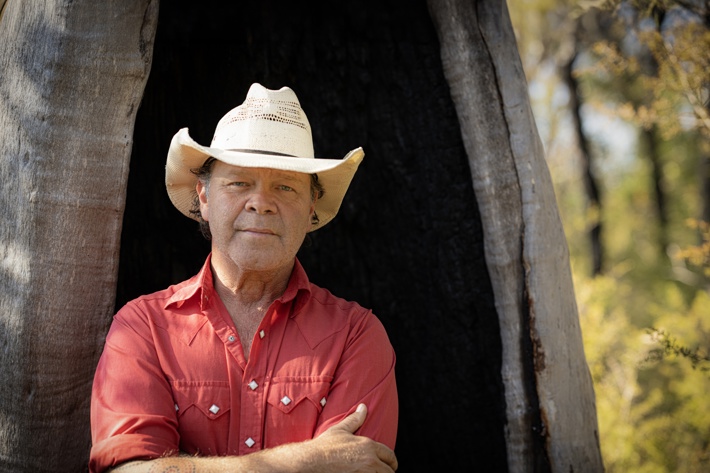 A contemplative person in a cowboy hat and red shirt stands beside a tree, gazing into the distance with a serene natural backdrop.