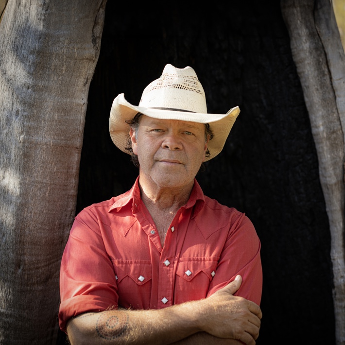 A contemplative person in a cowboy hat and red shirt stands beside a tree, gazing into the distance with a serene natural backdrop.