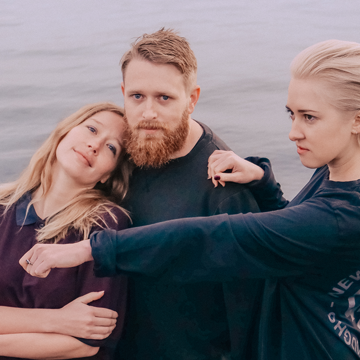 Three band members standing by the water, embracing with serene expressions