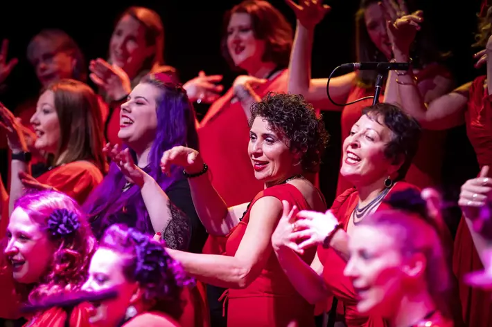 A choir group performs on stage wearing coordinated red outfits. They are positioned closely together, with arms raised and expressive hand gestures, suggesting a dynamic and engaging musical performance. The scene is brightly lit with pink and purple stage lighting, creating a vibrant atmosphere against a dark background.