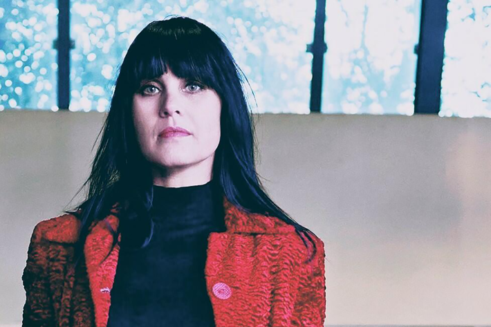 A person is standing indoors wearing a textured red coat with large buttons over a black high-neck top and black trousers. Hands are in the coat pockets, and long dark hair falls over the shoulders. The background shows a light-coloured wall and large windows with a view of trees outside.