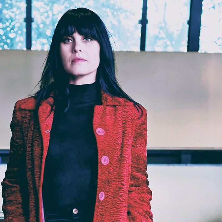 A person is standing indoors wearing a textured red coat with large buttons over a black high-neck top and black trousers. Hands are in the coat pockets, and long dark hair falls over the shoulders. The background shows a light-coloured wall and large windows with a view of trees outside.