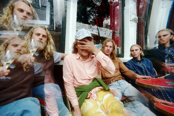 A group of people sitting closely together at an outdoor café table in front of a window with newspapers displayed inside. The setting includes red tables and chairs, and the individuals are dressed in casual clothing such as checked shirts, jeans, and layered outfits. One person has a green jumper tied around the waist, and another wears a cap. The image has a slightly distorted, double-exposure effect, adding a creative and dynamic feel.