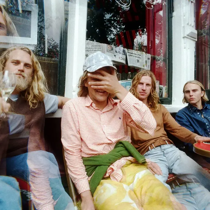 A group of people sitting closely together at an outdoor café table in front of a window with newspapers displayed inside. The setting includes red tables and chairs, and the individuals are dressed in casual clothing such as checked shirts, jeans, and layered outfits. One person has a green jumper tied around the waist, and another wears a cap. The image has a slightly distorted, double-exposure effect, adding a creative and dynamic feel.