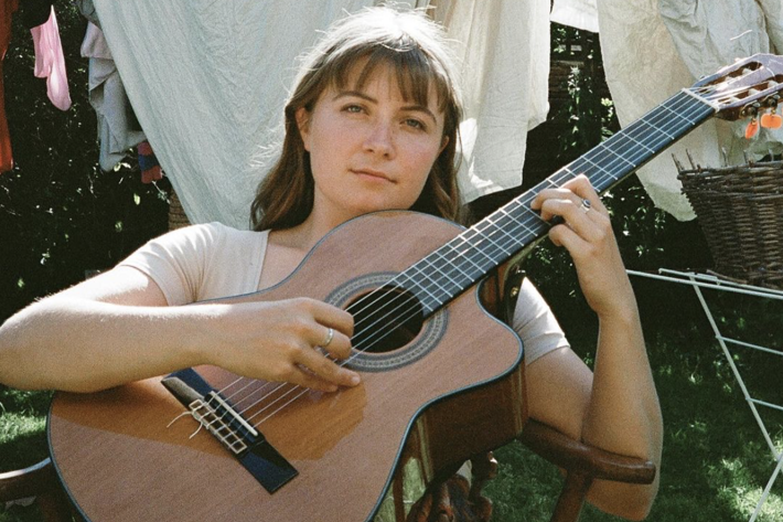 Jamie stares directly towards the camera sitting in a backyard on a wooden chair with a guitar held in front of her