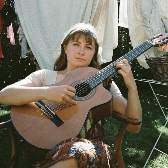 Jamie stares directly towards the camera sitting in a backyard on a wooden chair with a guitar held in front of her
