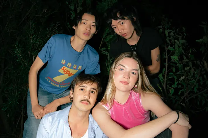Four people are gathered outdoors at night, sitting and leaning on a raised garden bed surrounded by greenery. One person in front wears a light blue shirt over a white top with jeans, while another beside them wears a bright pink sleeveless top and pinstriped trousers. Behind them, one person wears a blue graphic T-shirt and jeans, and another wears a black top. The scene is dimly lit, with plants and darkness in the background.