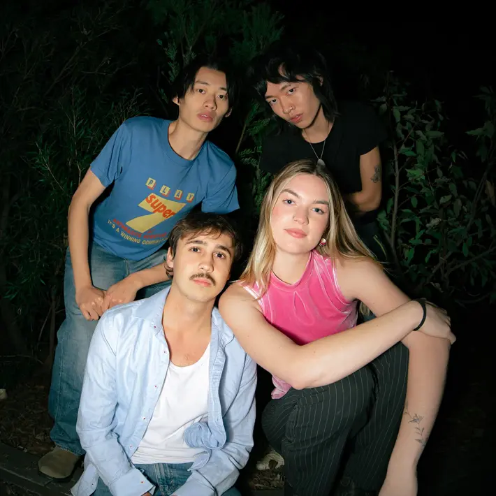 Four people are gathered outdoors at night, sitting and leaning on a raised garden bed surrounded by greenery. One person in front wears a light blue shirt over a white top with jeans, while another beside them wears a bright pink sleeveless top and pinstriped trousers. Behind them, one person wears a blue graphic T-shirt and jeans, and another wears a black top. The scene is dimly lit, with plants and darkness in the background.