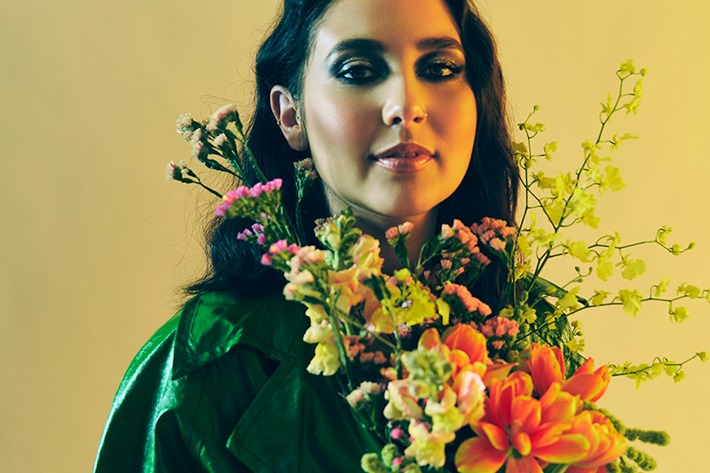 A person in a green coat holding a vibrant bouquet of flowers against a yellow background