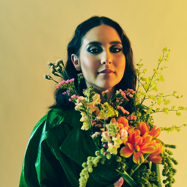 A person in a green coat holding a vibrant bouquet of flowers against a yellow background