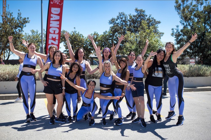 A group of people in athletic clothing pose energetically in an outdoor setting near a "DANCEZONE" banner.