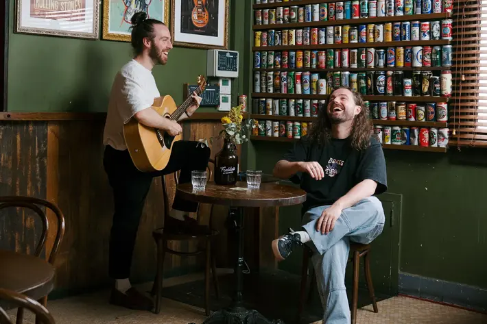 Two people sit and stand in a cosy pub-style room with green walls and wooden accents. One person plays an acoustic guitar while the other sits at a small round table with two glasses of water and a vase of flowers. Behind them, a large wall display features rows of colourful drink cans, and framed posters hang above. The atmosphere is warm and relaxed.
