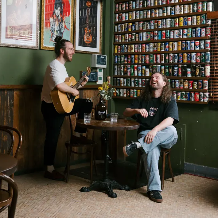 Two people sit and stand in a cosy pub-style room with green walls and wooden accents. One person plays an acoustic guitar while the other sits at a small round table with two glasses of water and a vase of flowers. Behind them, a large wall display features rows of colourful drink cans, and framed posters hang above. The atmosphere is warm and relaxed.