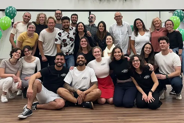 A group of people pose together in a dance studio with wooden flooring and a mirrored wall. Green balloons are visible on both sides, and a decorative arrangement of greenery hangs above the mirror. The group is casually dressed, with some wearing branded shirts, and they are arranged in two rows, suggesting a celebratory or social event related to dance.