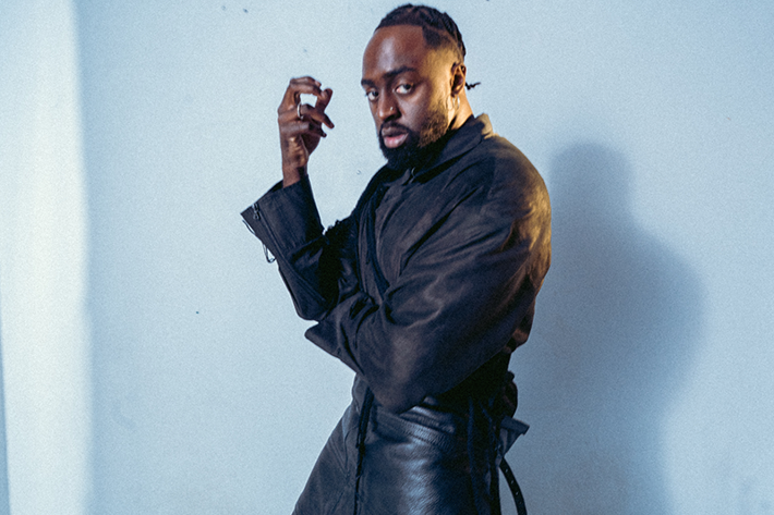 Person in a dark leather outfit striking a confident pose in a minimalist studio setting.