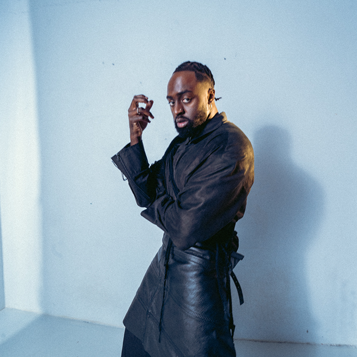 Person in a dark leather outfit striking a confident pose in a minimalist studio setting.