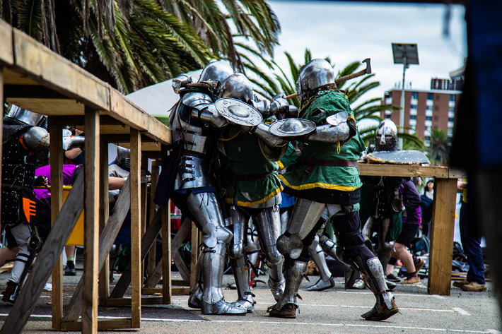 People dressed in full medieval-style armor are engaged in a mock combat or reenactment, pushing against each other with shields and swords near wooden barriers outdoors.