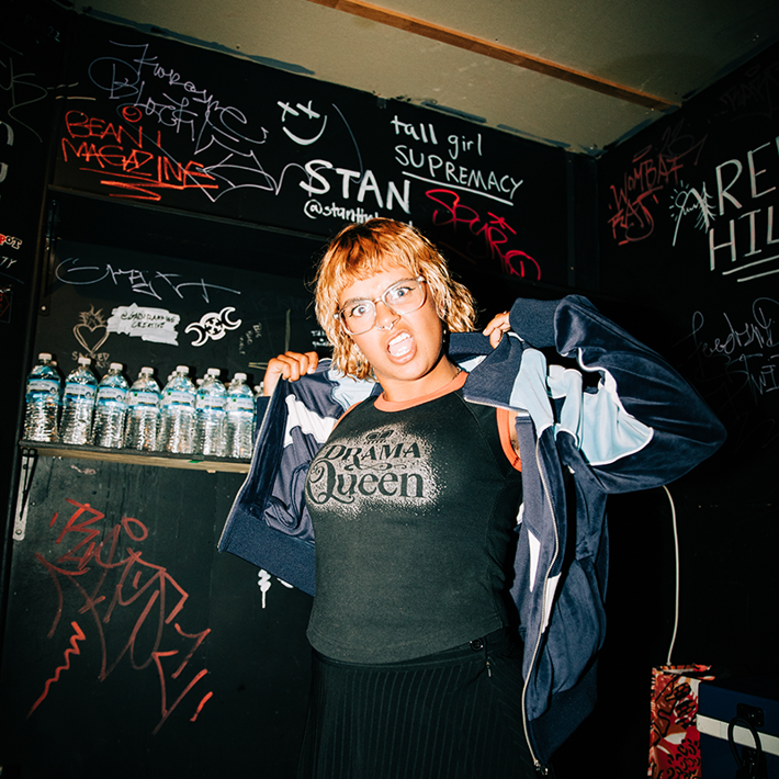 The image shows Bella, standing in front of a wall covered in graffiti. They are making a dramatic facial expression and wearing a t-shirt that says "Drama Queen".