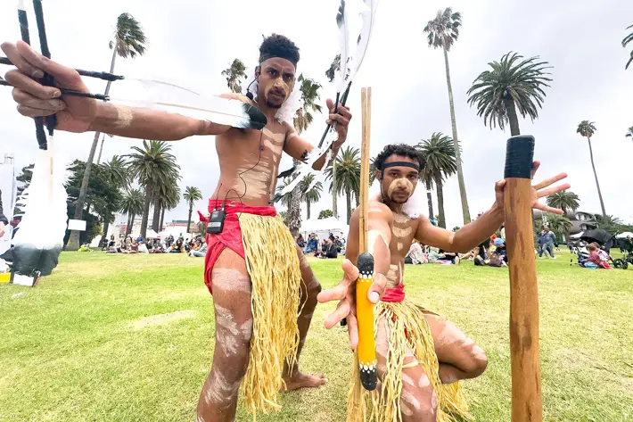 Two performers are engaged in a traditional cultural dance on a grassy outdoor area surrounded by palm trees. They are wearing grass skirts and red waistbands, with white body paint patterns on their skin. Each performer holds traditional items, including decorated sticks and feathers, and their dynamic poses suggest movement and storytelling. In the background, a crowd is seated, watching the performance, creating a vibrant community atmosphere.