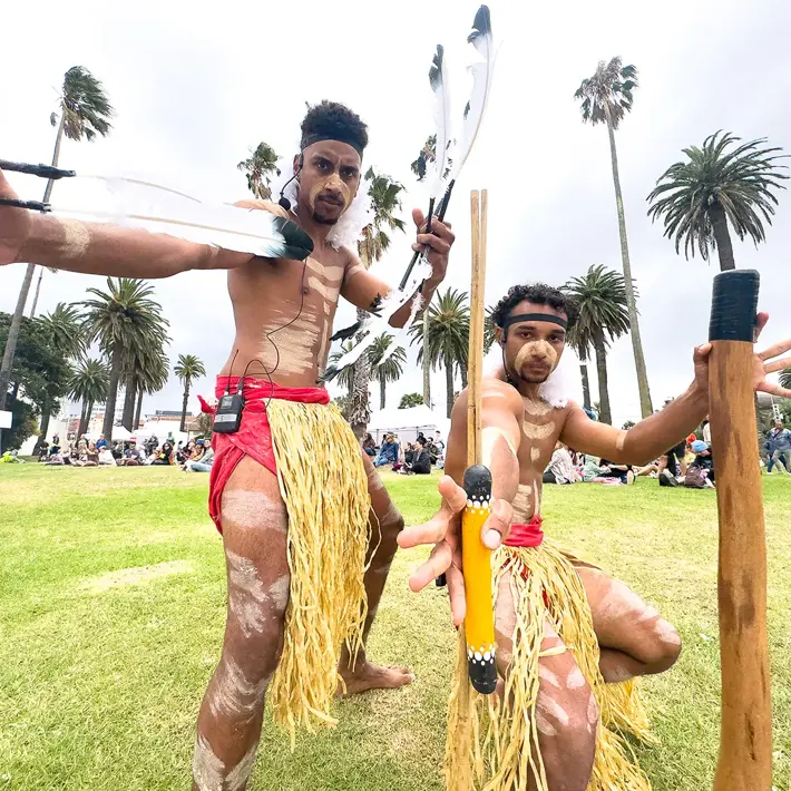 Two performers are engaged in a traditional cultural dance on a grassy outdoor area surrounded by palm trees. They are wearing grass skirts and red waistbands, with white body paint patterns on their skin. Each performer holds traditional items, including decorated sticks and feathers, and their dynamic poses suggest movement and storytelling. In the background, a crowd is seated, watching the performance, creating a vibrant community atmosphere.