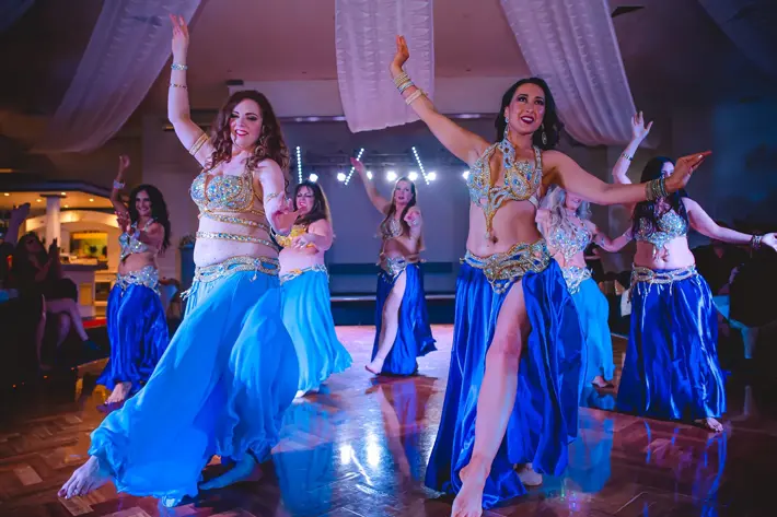 A group of performers are dancing on a polished wooden floor under soft purple and blue stage lighting. They wear coordinated costumes featuring blue flowing skirts and ornate gold and silver embellished tops, accessorised with matching jewellery. Their arms are raised in expressive poses, and the background includes draped white fabric and bright spotlights, creating an elegant and vibrant performance setting.