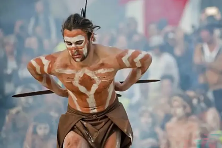 A person performing a traditional cultural dance outdoors, wearing a brown garment and body paint in white patterns across the chest, arms, and legs. The dancer holds two wooden sticks and has hair adorned with decorative elements. A large crowd is visible in the background, with smoke and banners adding to the ceremonial atmosphere.