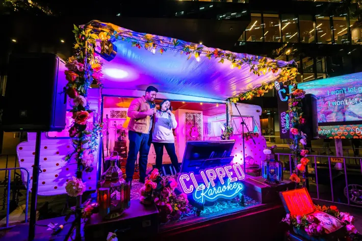 A vibrant outdoor karaoke setup features a decorated stage with colourful flowers, lanterns, and a neon sign reading “CLIPPED Karaoke.” Two people stand on the stage holding microphones, performing in front of a screen displaying song lyrics. The scene is illuminated with bright pink, purple, and blue lighting, creating a lively and festive atmosphere against a backdrop of modern buildings.