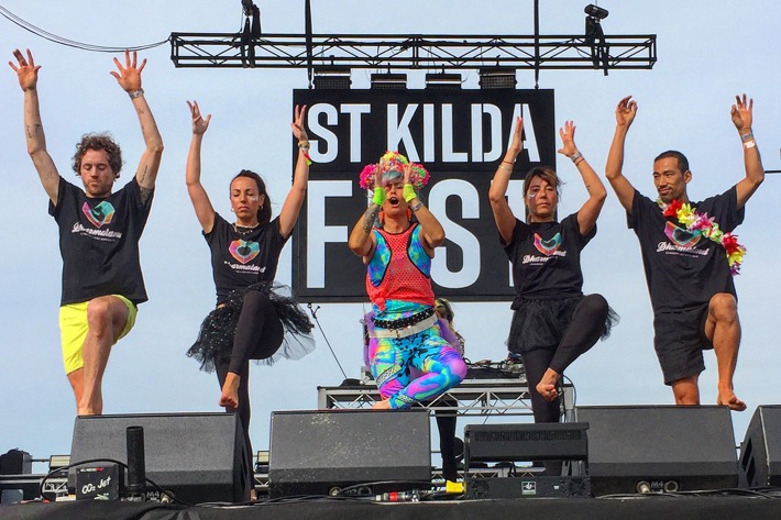 Five people standing in a line on the St Kilda Festival stage. They are all doing a yoga post with one leg and both arms raised. The person in the middle is wearing brightly coloured clothing and the rest are wearing black.
