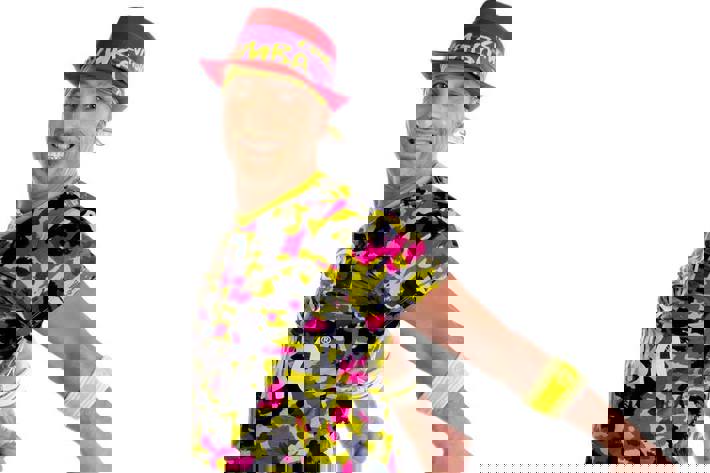Man jumping up with his arms back while wearing a red fedora, fluoro yellow leggings and a yellow, black and pink camouflage top all branded with the word Zumba.