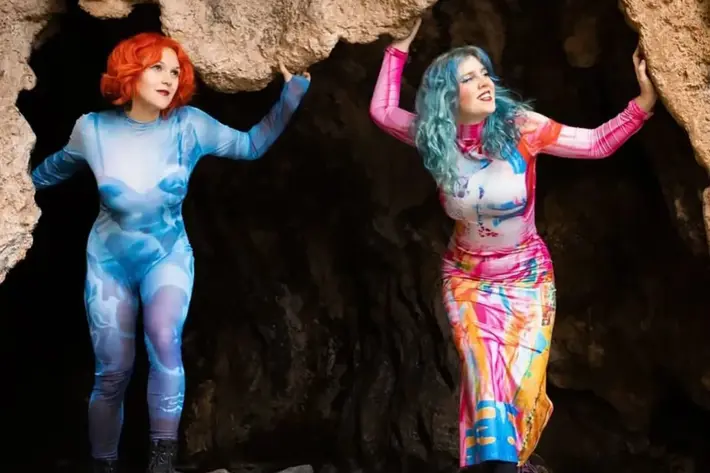 Credit: photographer is Makiko Ryujin, the image was taken on the Mornington Peninsula in a cave near Flinders Beach. Two people stand at the entrance of a rocky cave, each wearing vibrant, artistic outfits. One outfit features shades of blue with abstract patterns, while the other displays a mix of bright colours including pink, yellow, and blue. Both are holding onto the cave’s rough, textured edges, with rocky ground beneath them.