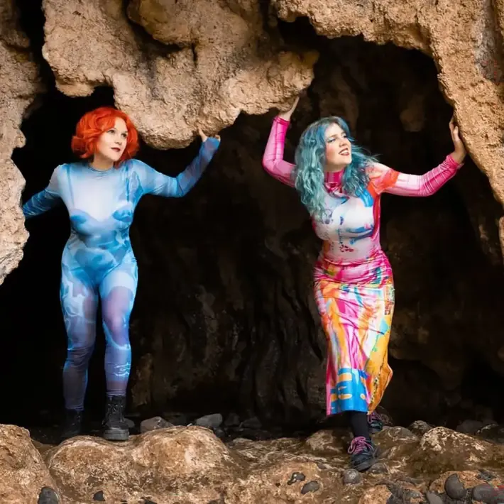 Credit: photographer is Makiko Ryujin, the image was taken on the Mornington Peninsula in a cave near Flinders Beach. Two people stand at the entrance of a rocky cave, each wearing vibrant, artistic outfits. One outfit features shades of blue with abstract patterns, while the other displays a mix of bright colours including pink, yellow, and blue. Both are holding onto the cave’s rough, textured edges, with rocky ground beneath them.