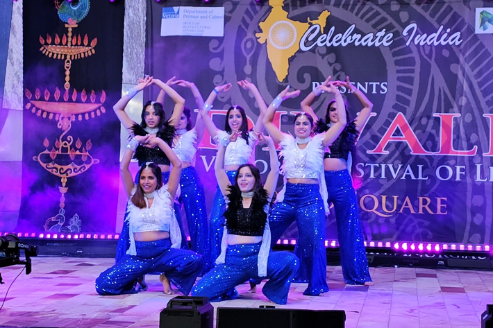 Seven women in Indian dancing attire, posing in a dance formation on stage.