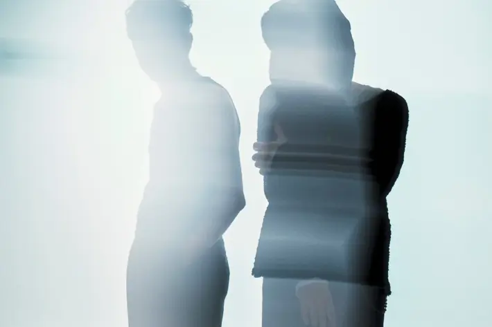Two silhouetted figures stand side by side in soft, diffused light. Their forms appear blurred and partially transparent, creating an abstract, atmospheric effect. One figure stands with hands in pockets while the other has arms loosely crossed. The background is pale and misty, giving the scene a calm, dreamlike quality.