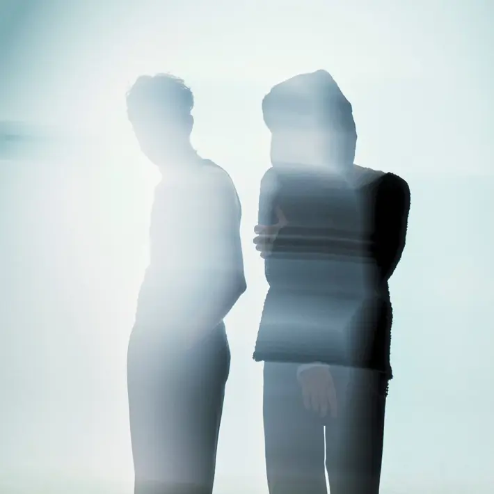 Two silhouetted figures stand side by side in soft, diffused light. Their forms appear blurred and partially transparent, creating an abstract, atmospheric effect. One figure stands with hands in pockets while the other has arms loosely crossed. The background is pale and misty, giving the scene a calm, dreamlike quality.