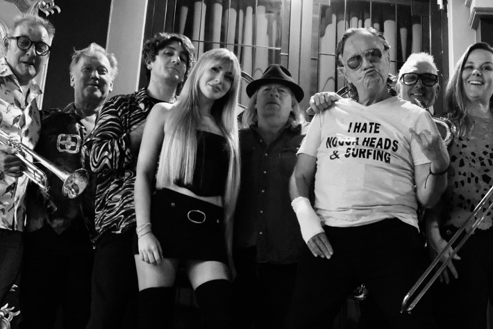 A black-and-white group photo featuring eight people standing closely together. Several hold musical instruments, including trumpets and a trombone. The group displays a mix of playful and confident expressions