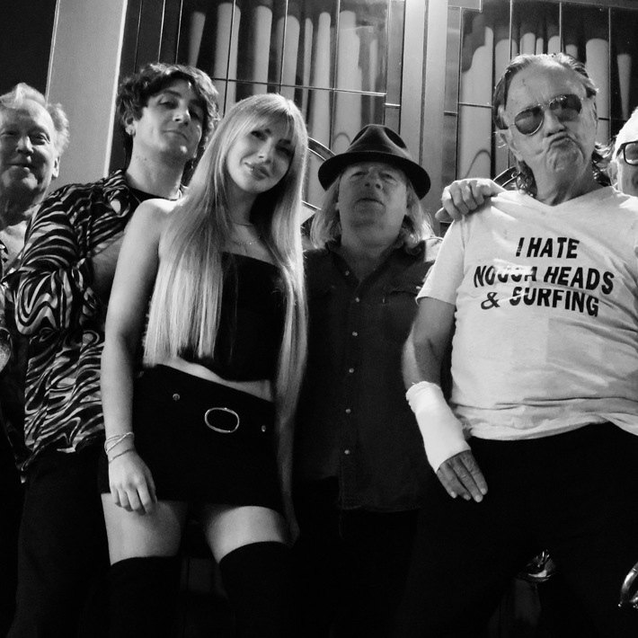 A black-and-white group photo featuring eight people standing closely together. Several hold musical instruments, including trumpets and a trombone. The group displays a mix of playful and confident expressions