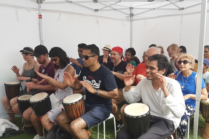 A diverse group of people sitting under a tent, each playing a hand drum with focused expressions.