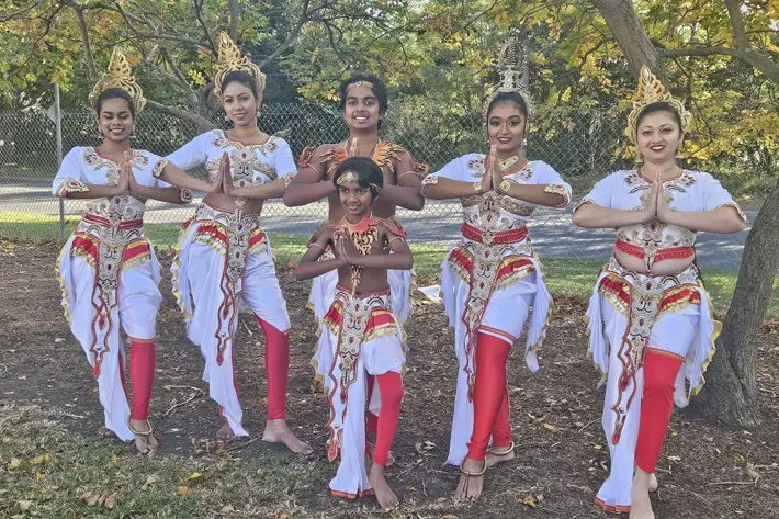 A group of performers pose outdoors under a tree, wearing traditional Sri Lankan dance costumes. The outfits feature ornate white garments with gold and red detailing, complemented by red leggings and decorative waistbands. Several performers wear elaborate gold headpieces, and all stand barefoot on grass with hands in a prayer position. The background includes a chain-link fence, trees with autumn leaves, and a paved road.