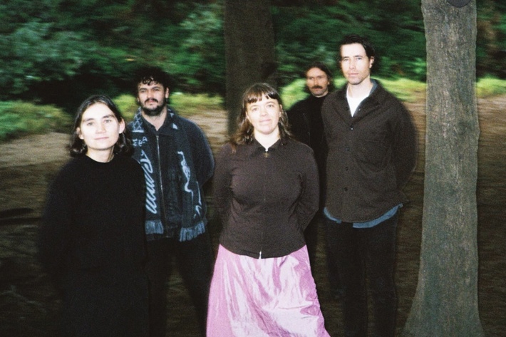Five members of the band EGGY stand in front of a blurred forest backdrop. The band members predominantly wear dark colours but a bright metallic pink skirt stands out in the centre. 
